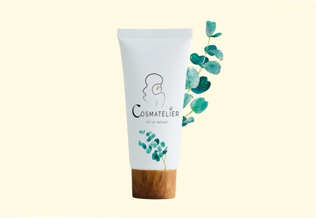Tube de crème de soin pour les cheveux cosmatelier. Sur fond beige pâle on fois un tube blanc avec un bouchon en bois. Sur le tube est dessiné le logo de Cosm'atelier, avec le visage doux d'une femme aux cheveux longs. Un élément végétal sort du tube.